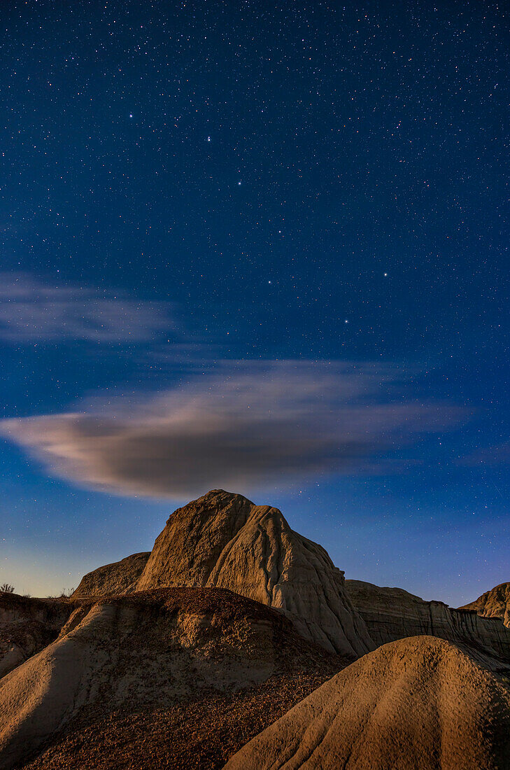 Der Große Wagen und Ursa Major über mondbeschienenen Formationen im Dinosaur Provincial Park, Alberta. Die Beleuchtung stammt vom zunehmenden Gibbous-Mond, der im Südwesten untergeht und so ein warmes "bronzefarbenes" Licht spendet.