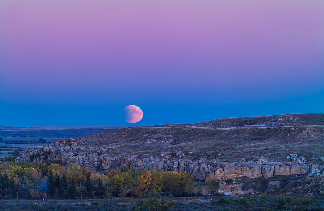 Der Vollmond geht in einer partiellen Finsternis über den Sandsteinformationen des Writing-on-Stone Provincial Park im Süden von Alberta am Abend des 27. September 2015 auf. In dieser Nacht fand eine totale Mondfinsternis statt, die sich in ihrer ersten partiellen Phase befand, als der Mond in dieser Nacht aufging. Das blaue Band am Horizont, das den Mond enthält, ist der Schatten der Erde auf unserer Atmosphäre, während der dunkle Biss, der aus der Mondscheibe herausgenommen wurde, der Schatten der Erde auf dem Mond ist. Das rosafarbene Band darüber ist der Gürtel der Venus.