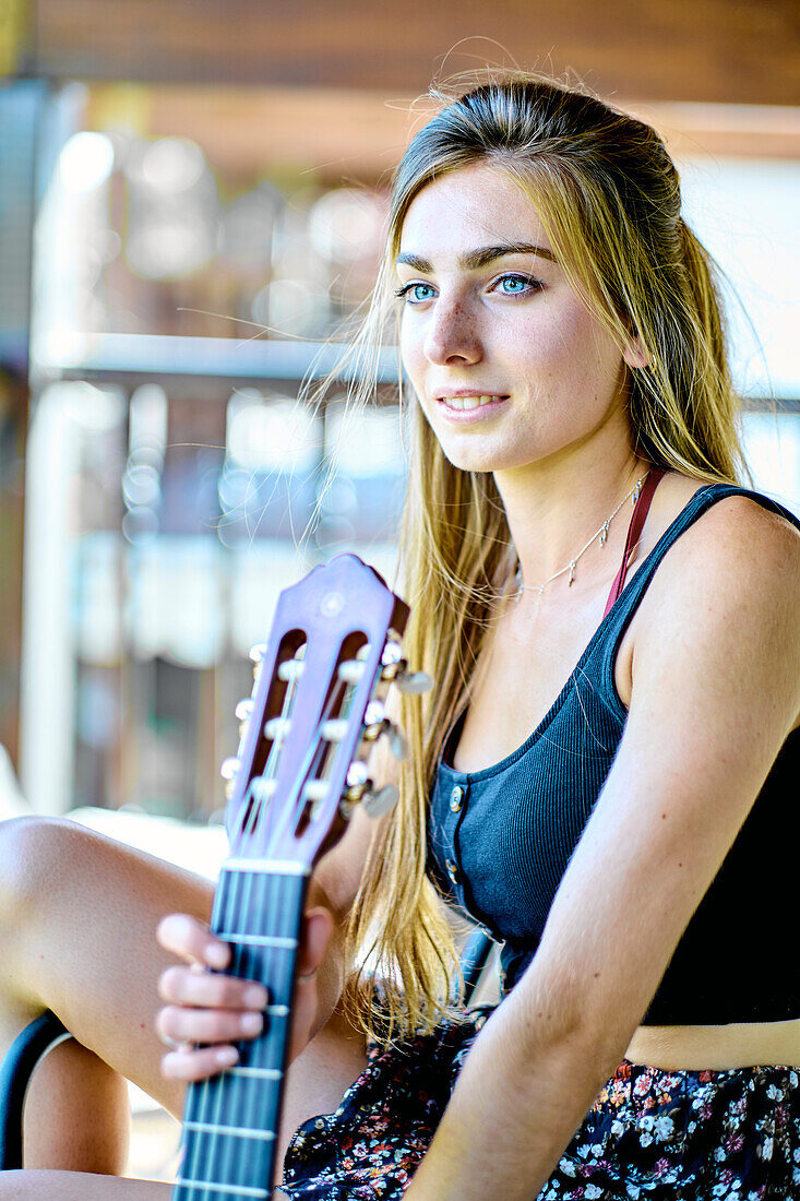 Portrait einer jungen schönen kaukasischen Frau in den 20ern mit langen Haaren und blauen Augen mit einer Gitarre auf der Veranda eines Landhauses. Lifestyle-Konzept.