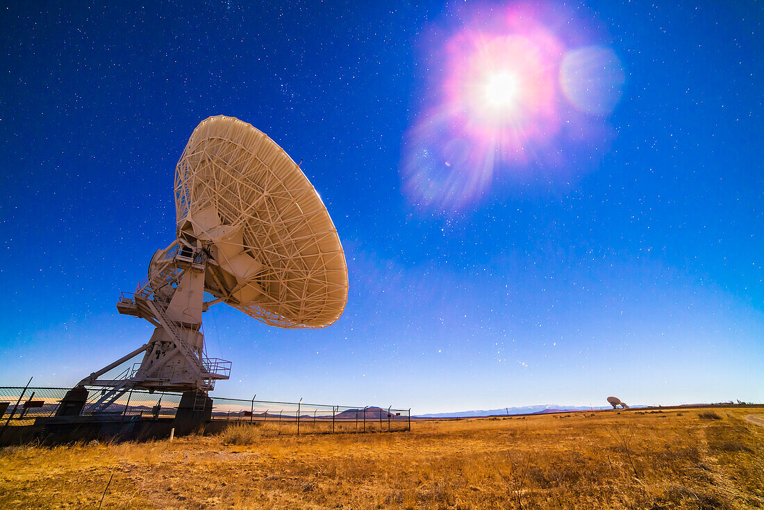 One of the 27 antennas of the Very Large Array (VLA) radio telescope complex in New Mexico (with others in the distance at lower right) illuminated by moonlight, on December 13, 2013, peak night for the Geminid meteor shower. A single exposure of 30 seconds with the Rokinon 14mm lens at f/2.8 and Canon 5D MkII at ISO 800. Orion is rising a lower centre. The Moon is the bright object at upper right. The Pleiades and Hyades are above centre.
