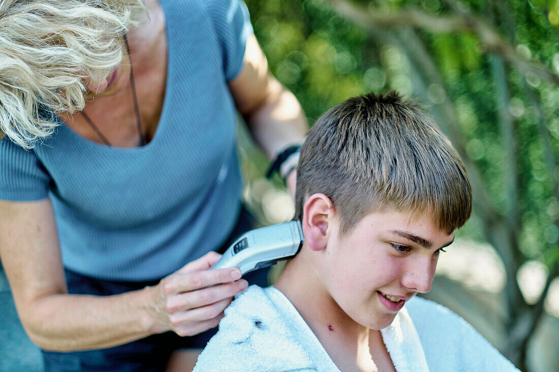 Porträt einer Mutter, die einem kleinen kaukasischen Jungen draußen im Garten die Haare schneidet. Lifestyle-Konzept.