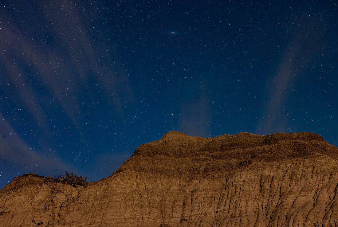 The Andromeda Galaxy (aka Messier 31) and the stars of Andromeda rising over moonlit formations at Dinosaur Provincial Park, Alberta. Illumination is from the waxing gibbous Moon, low in the southwest so it is providing a warm light. This is an example of a "deepscape" - a nightcape with a telephoto lens to also record a deep-sky object in the frame above the horizon. However, moonlight prevents the galaxy from showing up as well as it would in a dark sky.
