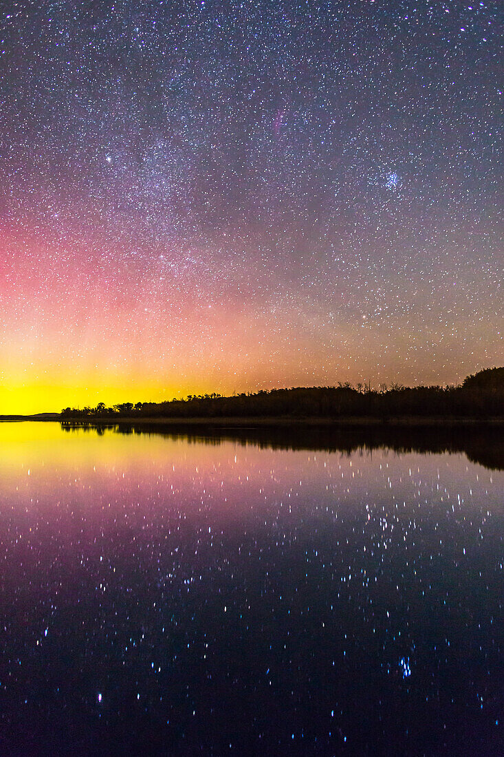 Die aufgehenden Herbststerne und Sternbilder über dem See im Police Outpost Provincial Park im Süden von Alberta, am 26. September 2016. Die Sterne von Auriga und Taurus gehen auf, darunter die Plejaden oben rechts. Capella ist der helle Stern oben rechts in der Mitte; Aldebaran steht unten rechts am Himmel. Beide spiegeln sich im stillen Wasser, zusammen mit den Sternhaufen Hyaden und Plejaden. Links ist ein leichtes Polarlicht zu sehen.