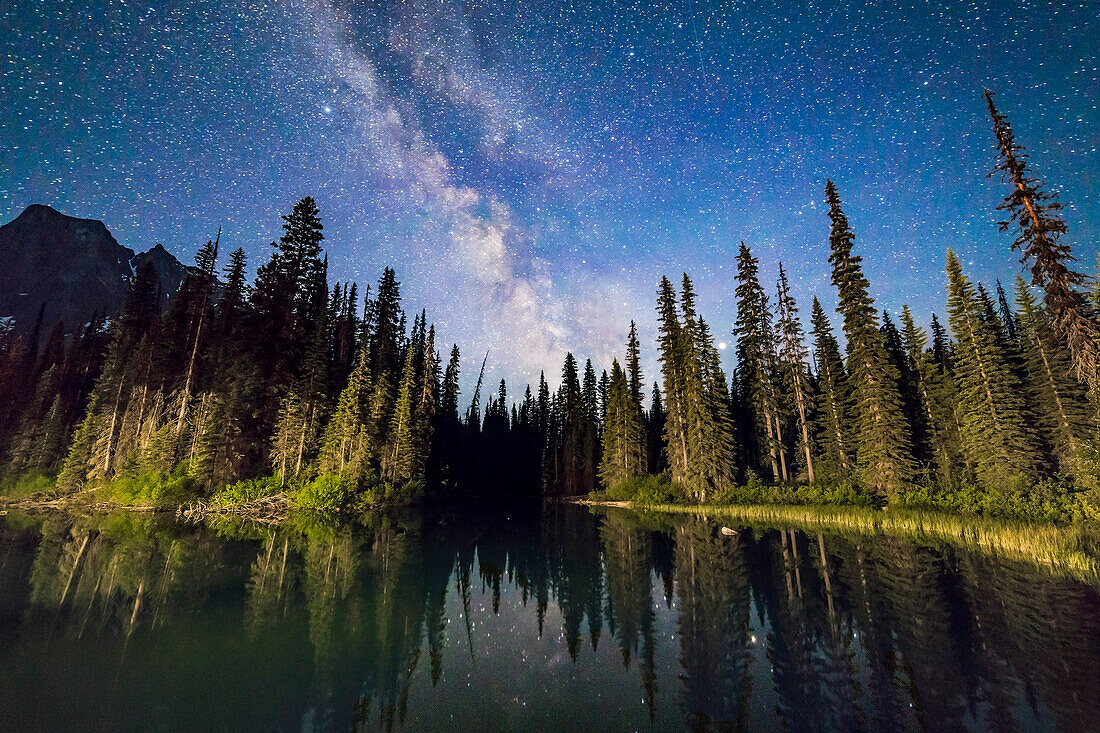 Die Milchstraße über dem Seitenteich am Emerald Lake, Yoho National Park, BC, von der Brücke zur Lodge. Die Lichter der Lodge erhellen die Bäume. Die ewige Dämmerung kurz vor der Sonnenwende (aufgenommen am 6. Juni 2016) lässt den Himmel tiefblau leuchten. Saturn ist das helle Objekt im Dunst, das rechts durch die Bäume scheint.