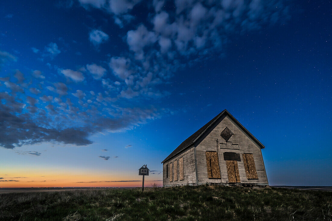 Das Liberty Schoolhouse von 1910, eine klassische Einraumschule der Pioniere, in der Prärie von Alberta unter dem Sternenhimmel in der Dämmerung einer Frühlingsnacht. Das Mondlicht des zunehmenden Mondes sorgt für die Beleuchtung.