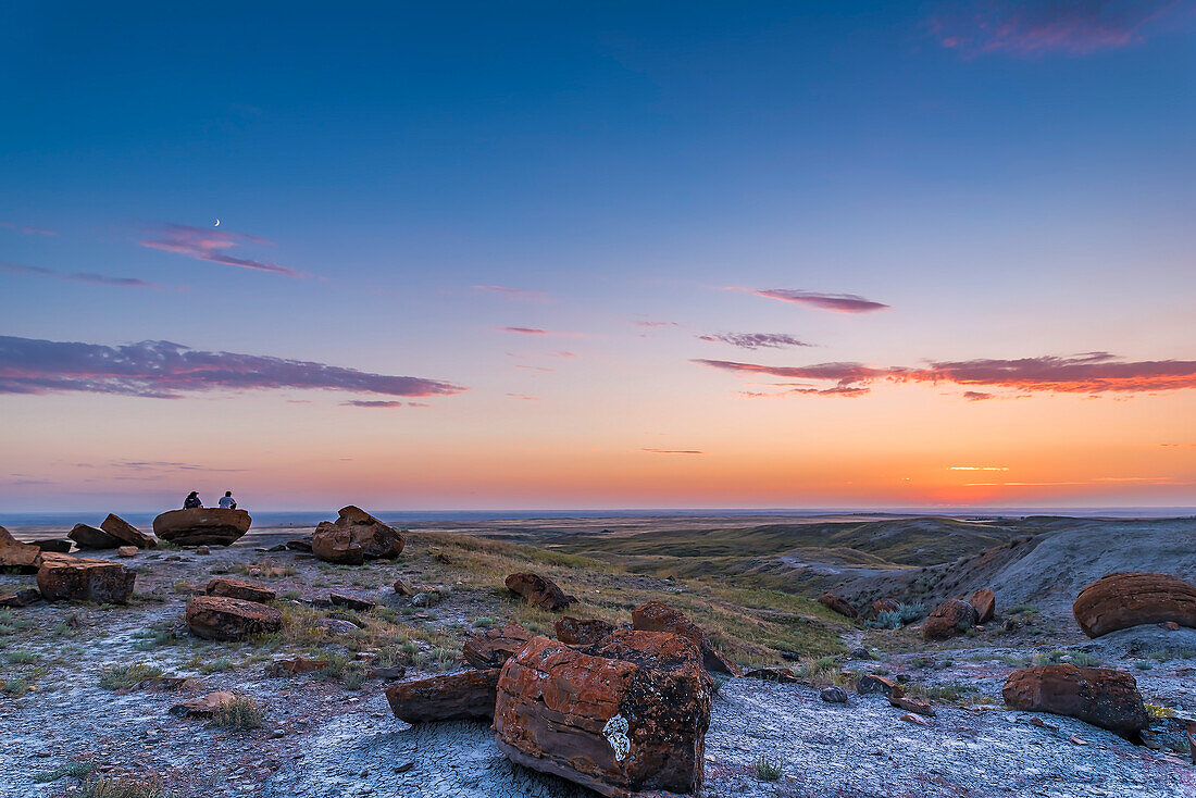 A pair of campers watch the sunset with the 4-day-old waxing Moon in the twilight sky above, at Red Rock Coulee in southeast Alberta.