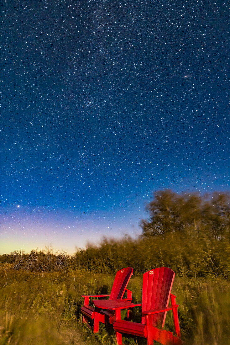 Die ikonischen roten Stühle von Parks Canada am Teich zur Beobachtung von Wasservögeln am Eingang des Waterton Lakes National Park, Alberta, Kanada, an einem mondbeschienenen Herbstabend, an dem die Sterne des Herbsthimmels im Nordosten aufgehen. Zu sehen sind Perseus, das W der Kassiopeia und der größte Teil von Andromeda, einschließlich der Andromedagalaxie oben rechts. Der helle Stern unten links ist Capella in Auriga.