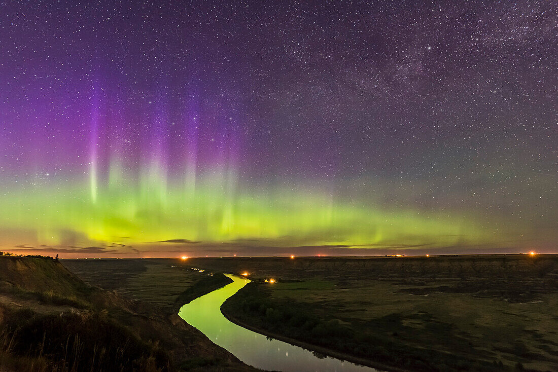 Das Nordlicht tanzt über dem Red Deer River und den Badlands in Süd-Alberta, vom Orkney Viewpoint mit Blick nach Norden über das Tal. Die Bleriot-Fähre ist in der Ferne bei den Lichtern zu sehen. Kassiopeia ist in die violetten Vorhänge eingebettet. Der Fluss reflektiert das Licht der Aurora.