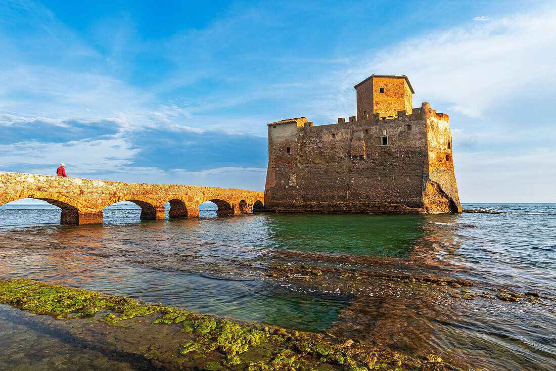 Man on the bridge walks to the fortified castle of Torre Astura in the water of Tyrrhenian Sea built on ruins of Roman villa, sunset time, Rome province, Latium (Lazio), Italy, Europe