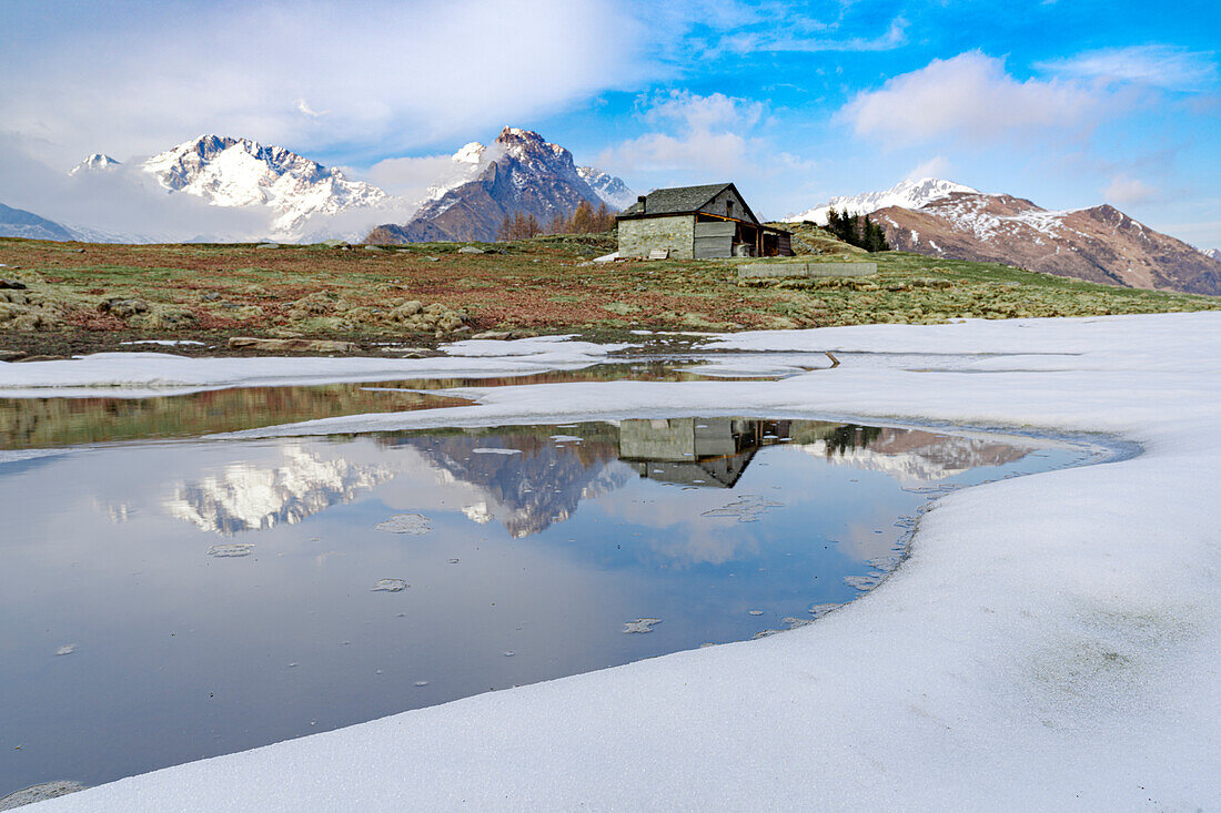 Berghütte am Ufer eines zugefrorenen Teiches während des Tauwetters, Scermendone Alp, Rätische Alpen, Valtellina, Lombardei, Italien, Europa