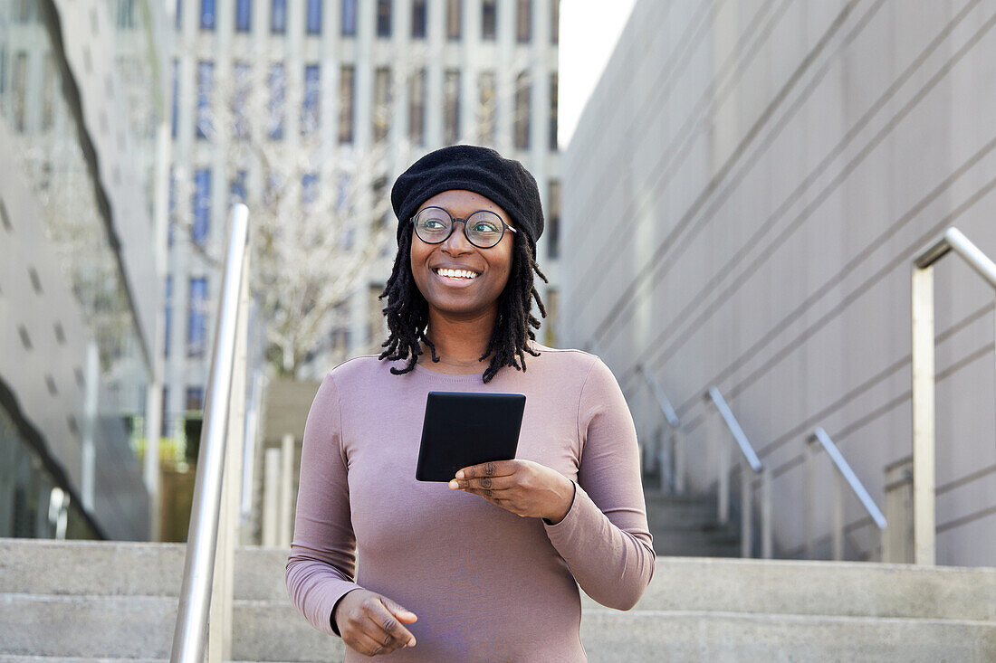 Lächelnde Frau mit Brille und Baskenmütze hält digitales Tablet in der Stadt