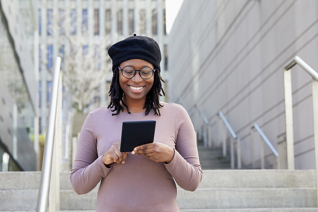 Smiling woman wearing eyeglasses and beret holding digital tablet in city