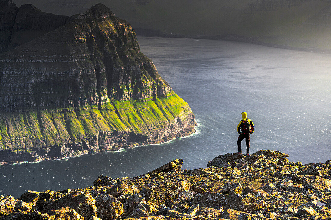 Ein Mann bewundert die majestätischen Klippen entlang eines Fjordes auf dem Gipfel eines Berges während einer Wanderung, Vidoy Island, Färöer Inseln, Dänemark, Europa