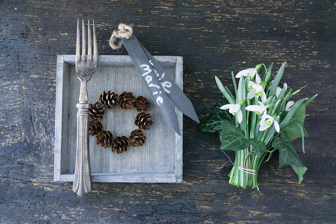 Holztablett mit Kranz aus Lärchenzapfen und Silbergabel, Strauß aus Schneeglöckchen (Galanthus) mit Efeublättern und Namensschildchen