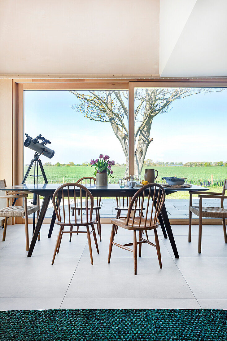 Dining area with wooden chairs and telescope in front of panoramic window with countryside view