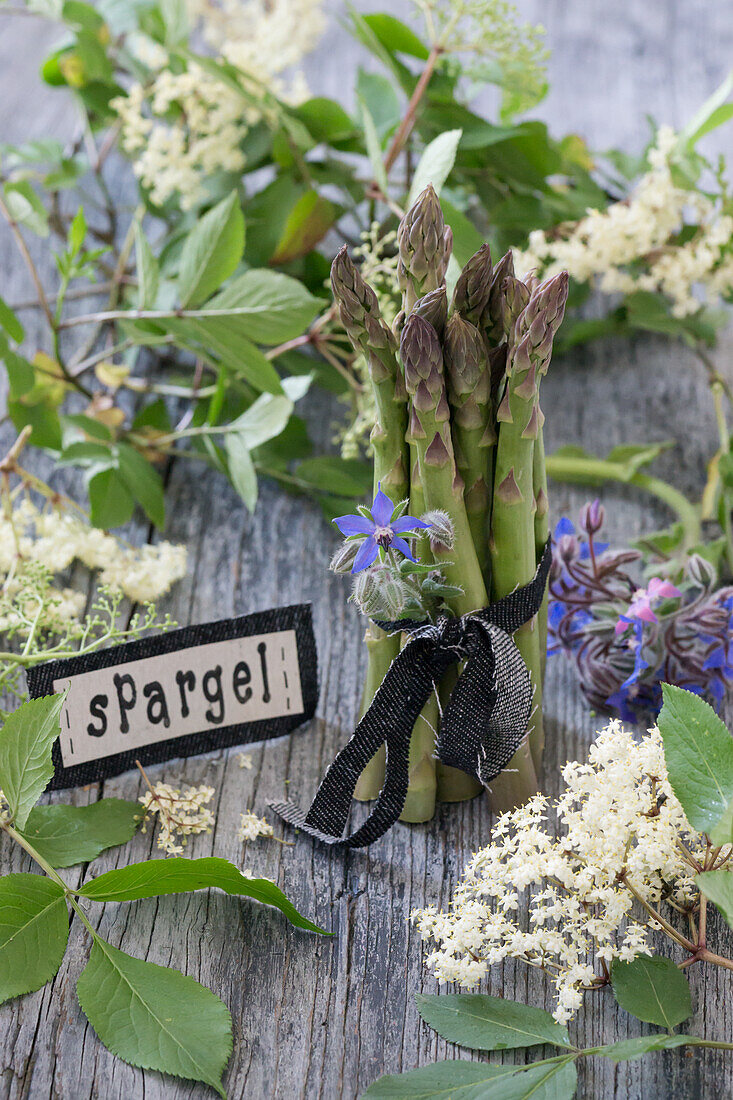 Bund grüner Spargel mit Holunderblüten (Sambucus Nigra), Borretschblüten (Borago officinalis) und Jeansband auf Holzuntergrund