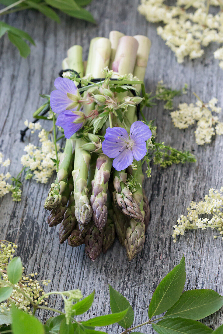 Bund grüner Spargel mit Storchenschnabel (Geranium) und Holunderblüten (Sambucus Nigra) auf Holzuntergrund