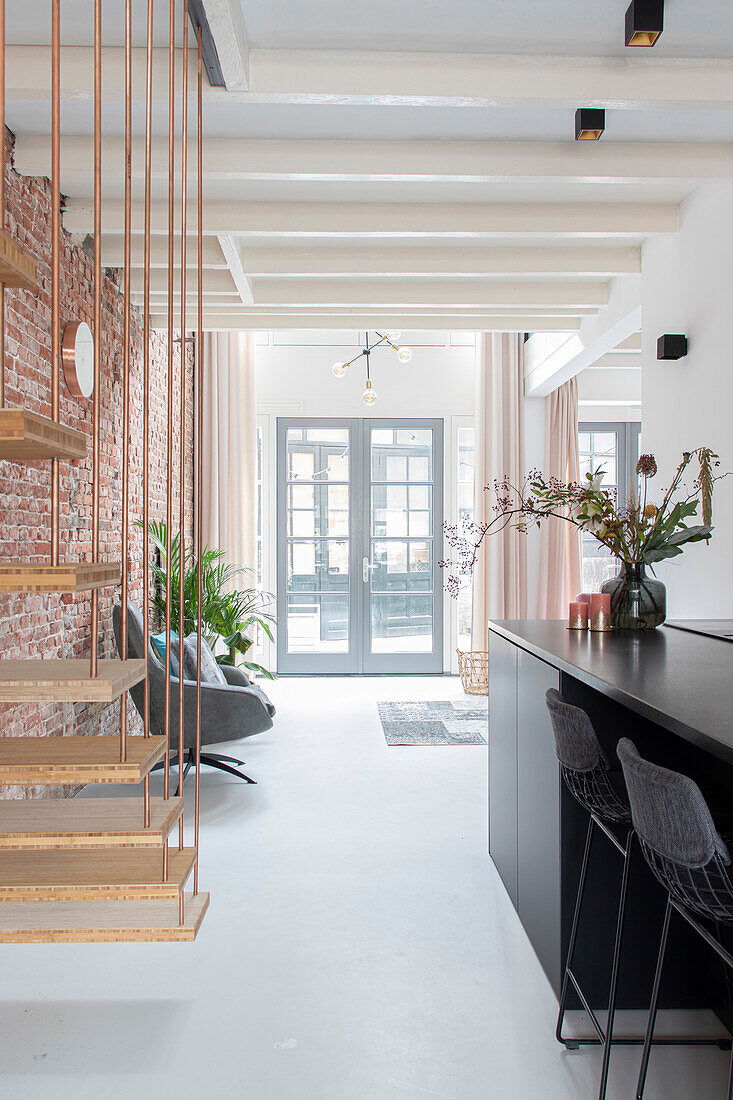 Open-plan living room with free-floating wooden staircase, brick wall and white-painted ceiling beams