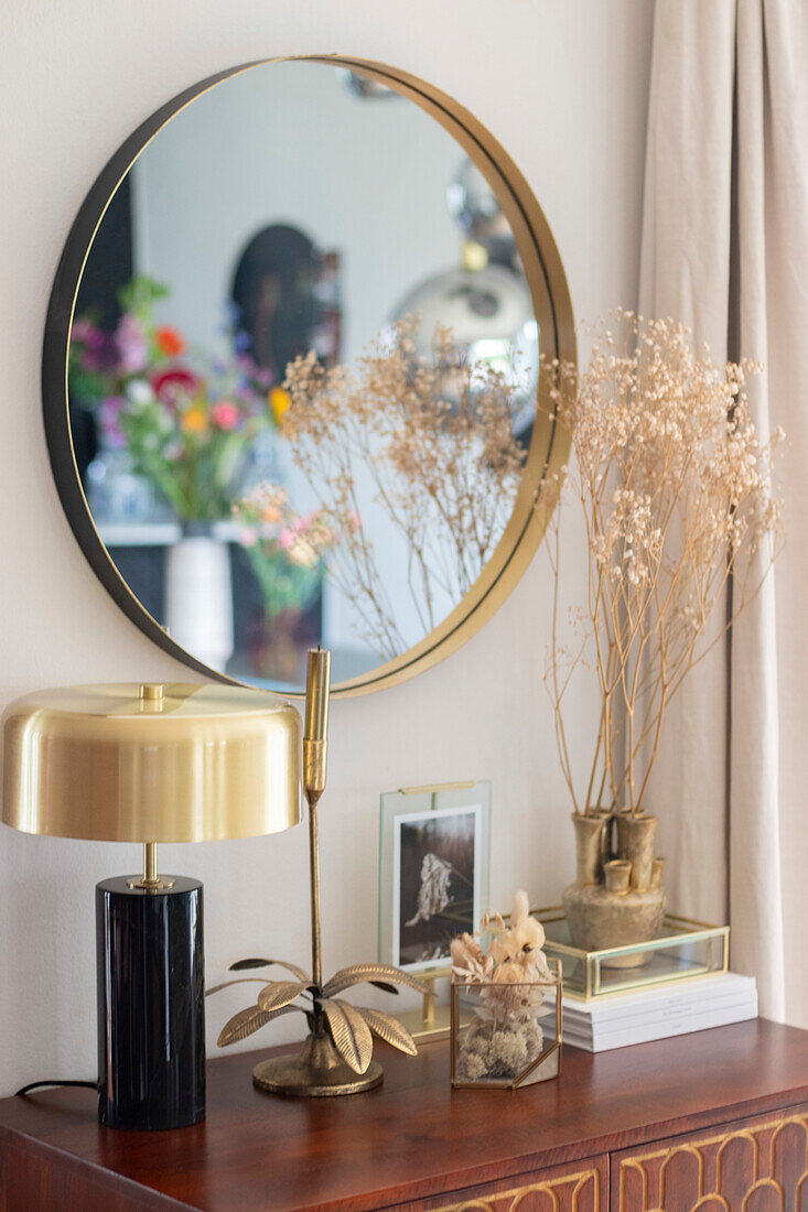 Round mirror with golden frame above decorated sideboard with dried flowers and golden table lamp