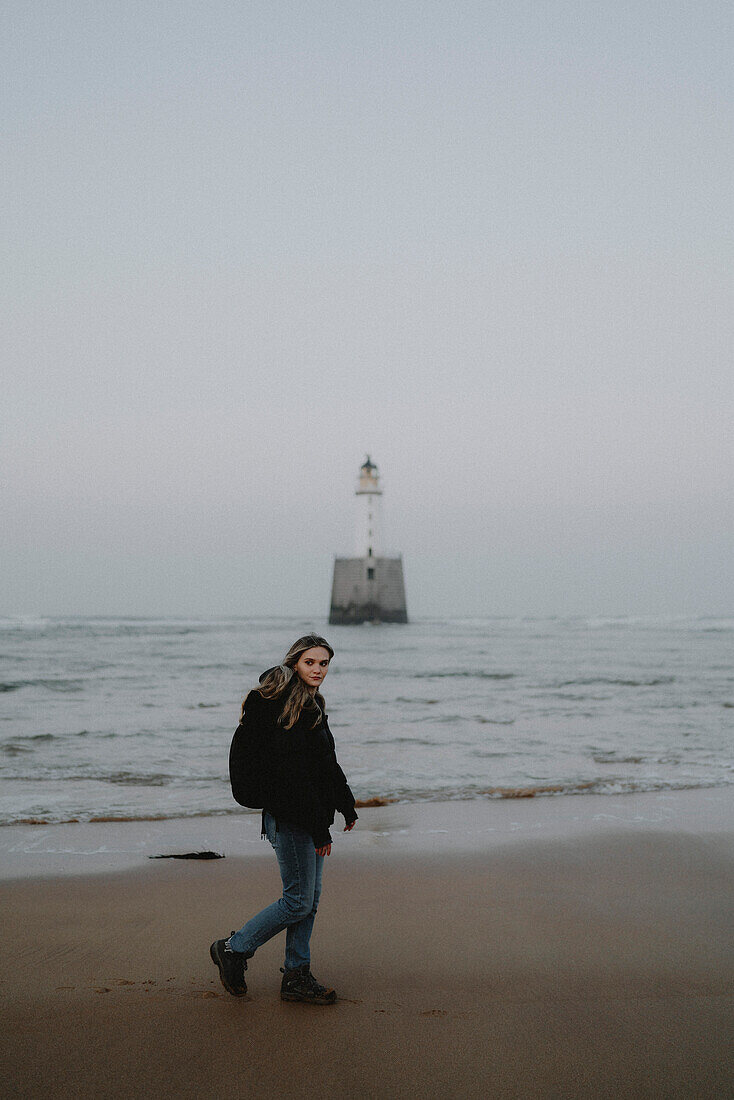 Portrait young woman walking past lighthouse on wet sand beach, Rattray, Aberdeenshire, Scotland\n