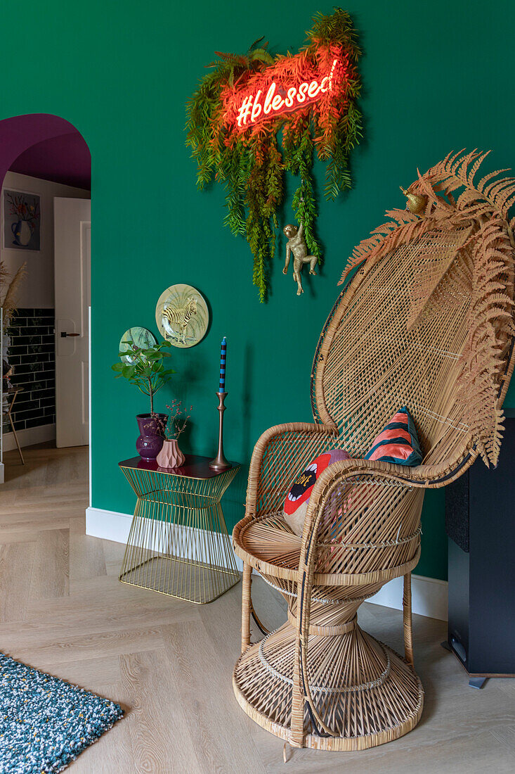 Rattan high-back armchair and golden side table in front of green wall with neon blessed sign