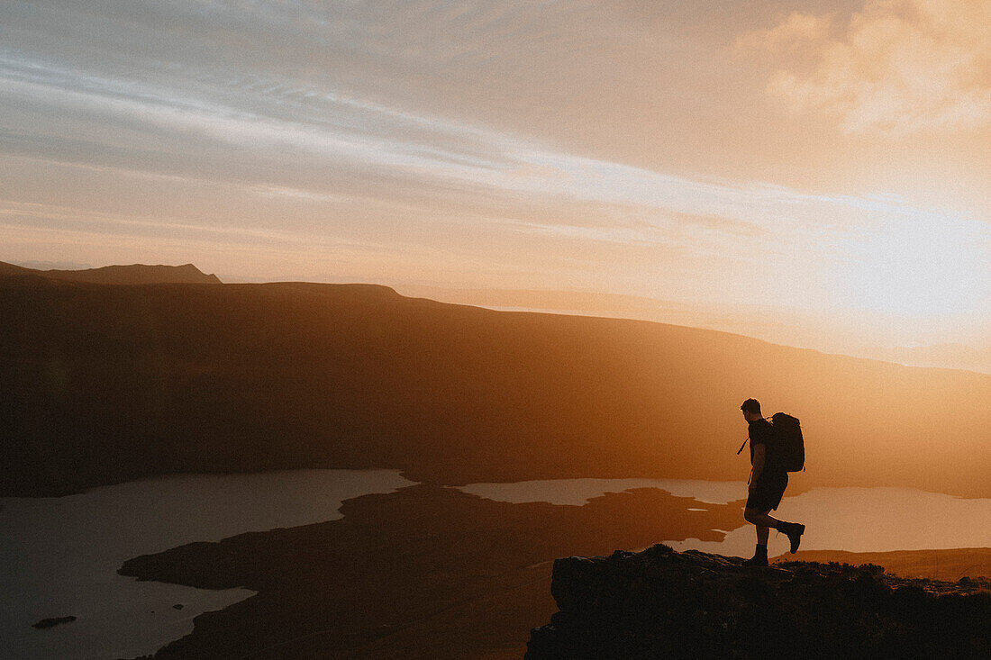 Silhouetted hiker on mountain overlooking idyllic sunset view, Assynt, Sutherland, Scotland\n