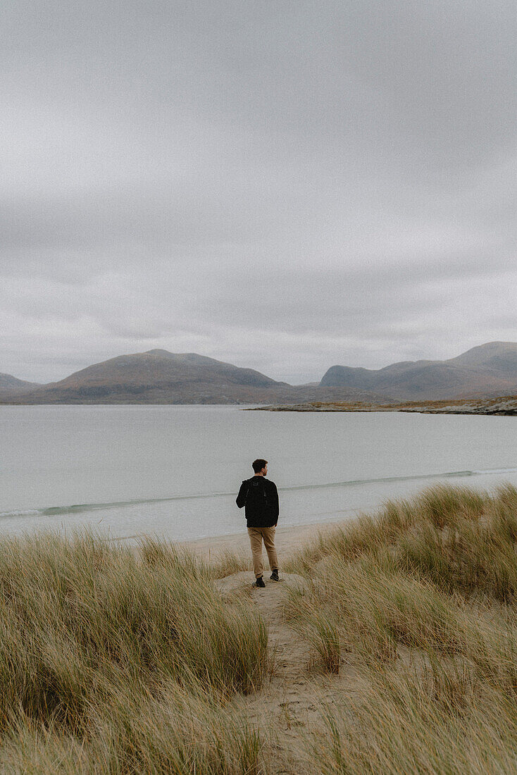 Male hiker standing among sandy beach grass at ocean, looking at mountain view, Scotland\n