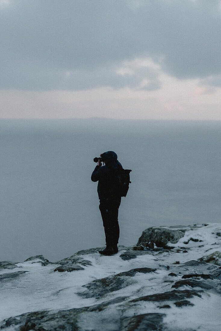 Fotograf beim Wandern und Fotografieren des Ozeans von den schneebedeckten Klippen aus, Neist Point, Isle of Skye, Schottland