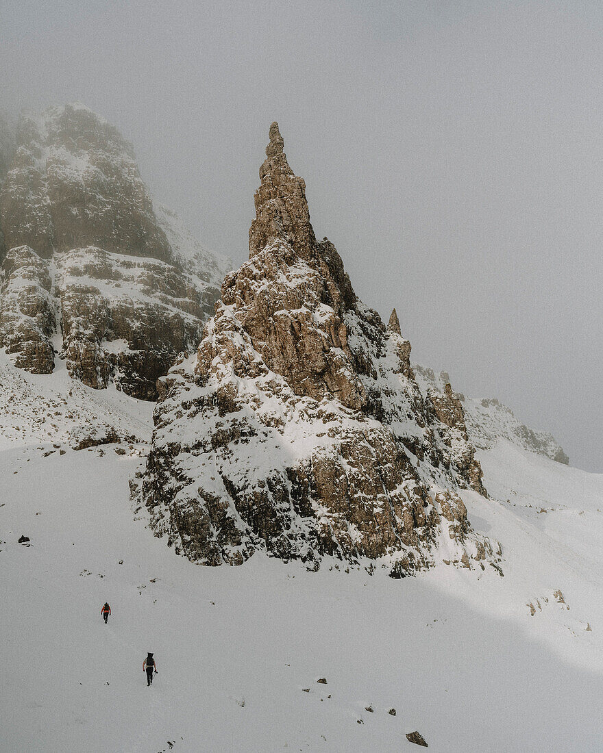 Hikers climbing mountain slope below snow covered rock formation, Old Man of Storr, Scotland\n