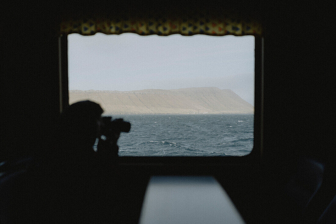 Silhouetted photographer with camera photographing ocean view from ferry, Sandur, Faroe Islands\n