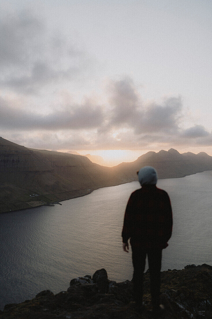 Mann auf dem Gipfel eines Hügels mit Blick auf den Sonnenuntergang über dem Fluss, Klakkur, Klaksvik, Färöer Inseln