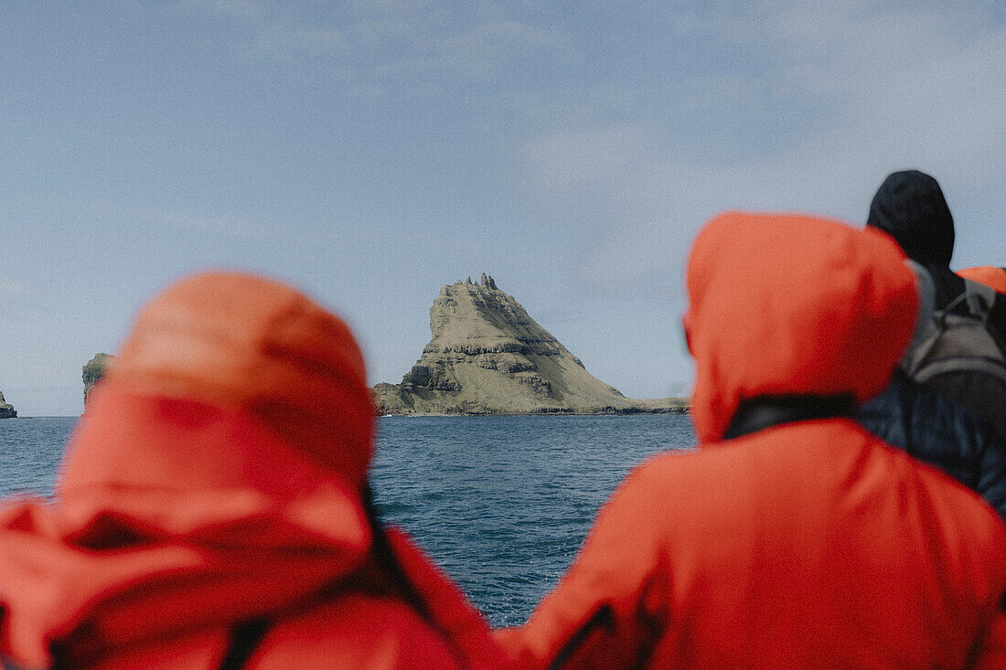 Tourists on ferry looking at rock formation over sunny ocean, Mykines, Faroe Islands\n