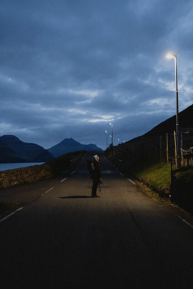 Man on dark road looking up at streetlamp, Kunoy, Faroe Islands\n