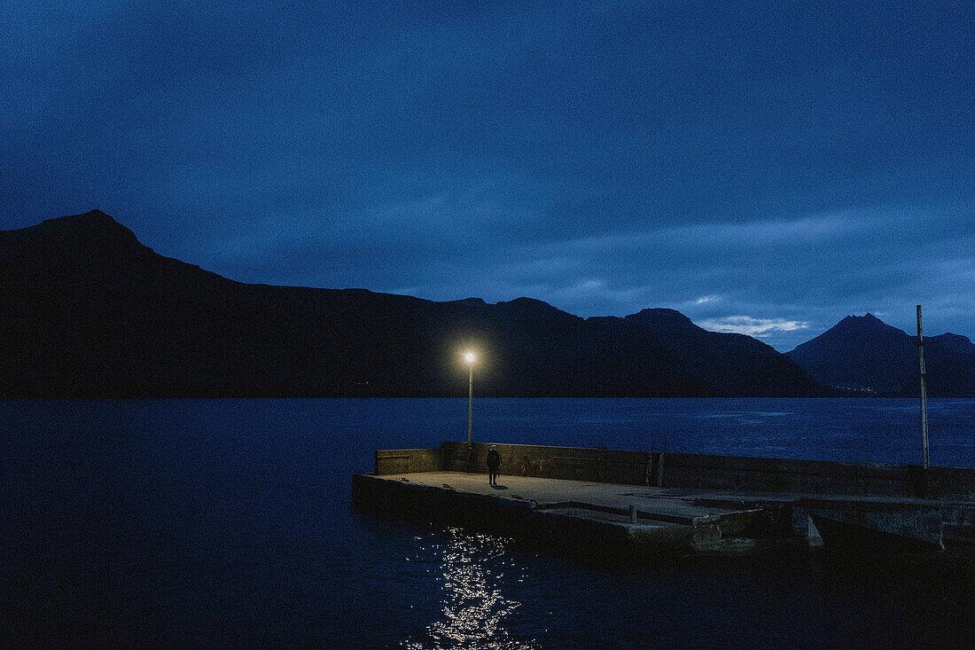 Mann im Dunkeln auf Bootssteg unter Straßenlaterne und dunkelblauem Nachthimmel, Kunoy, Färöer Inseln