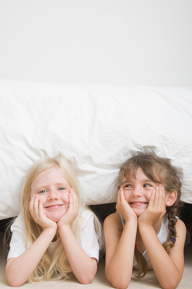 Zwei junge Mädchen mit auf die Hände gestütztem Kinn unter dem Bett lächelnd