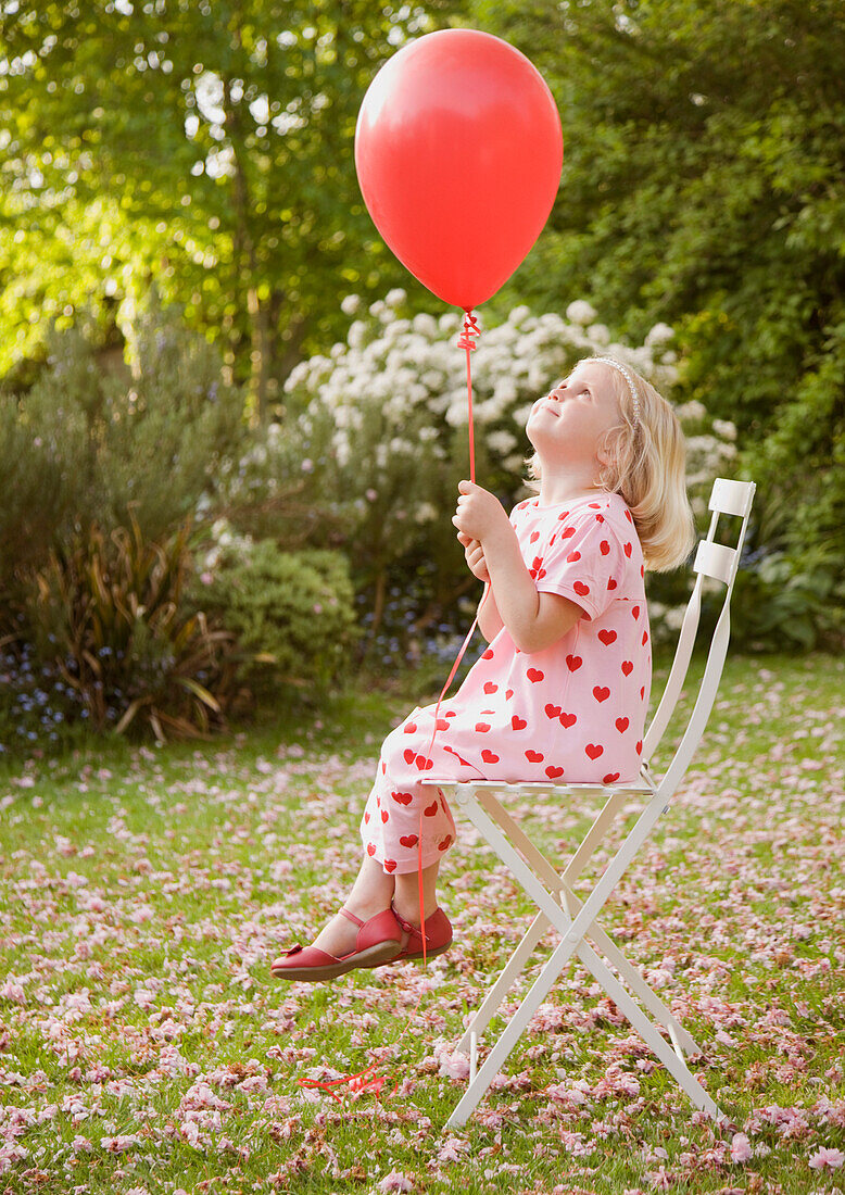 Young girl sitting on a chair in the garden holding a red balloon\n