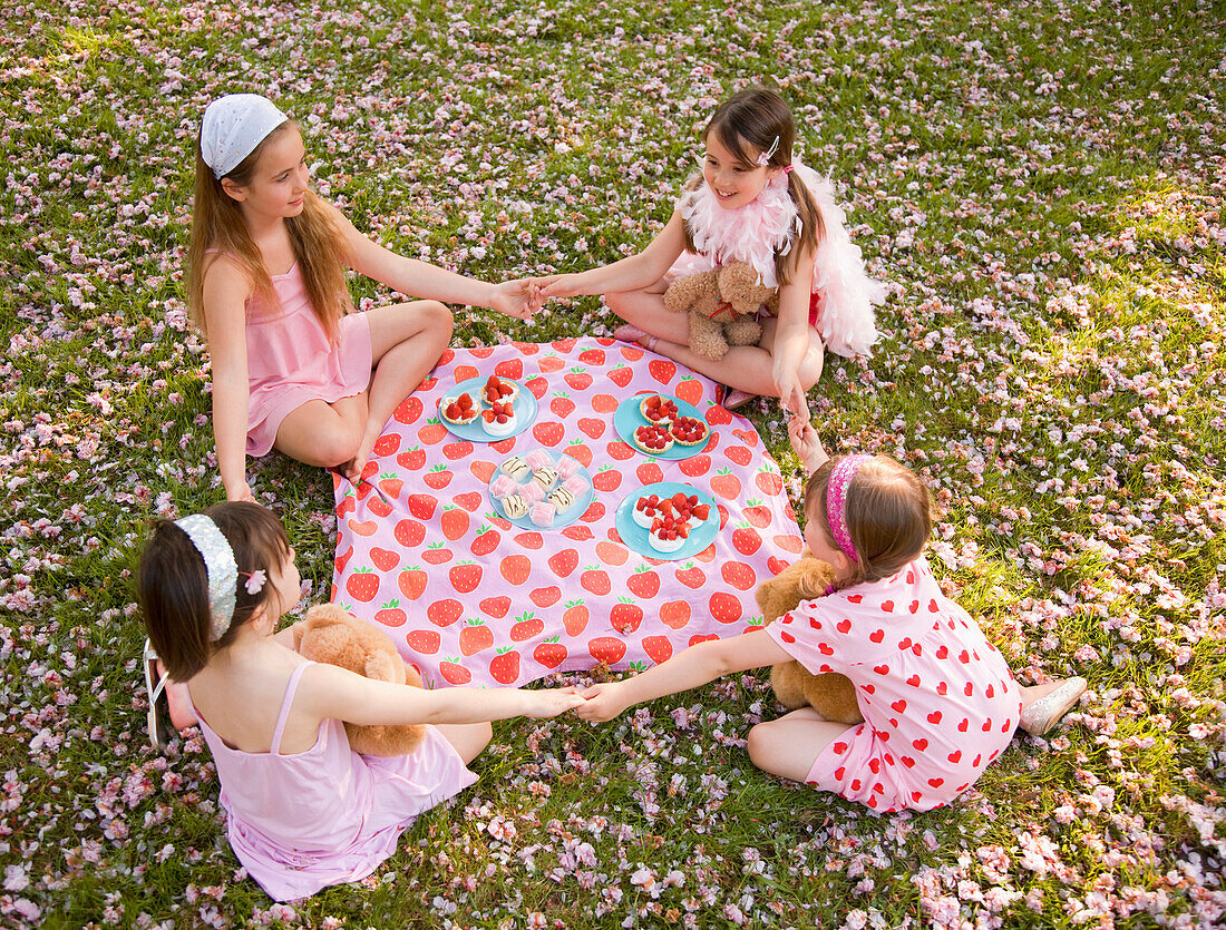 Four young girl sitting around a colorful blanket holding hands and having a party\n