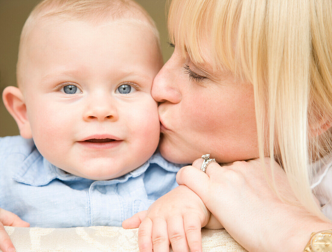 Close up of a smiling baby with his mother kissing him\n