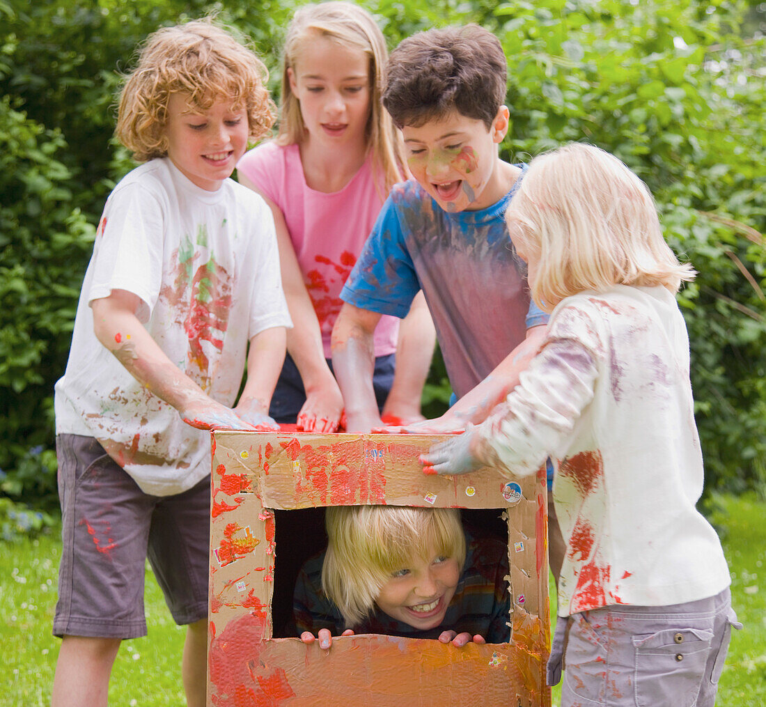 Kinder mit Aquarellfarbe bedeckt spielen im Garten, eines von ihnen steht in einem Pappkarton