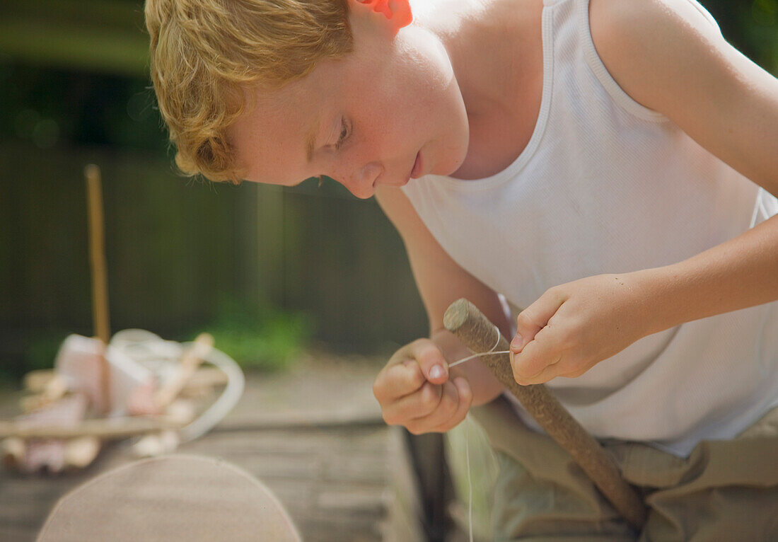 Young boy tying a knot with a string around a wooden stick\n