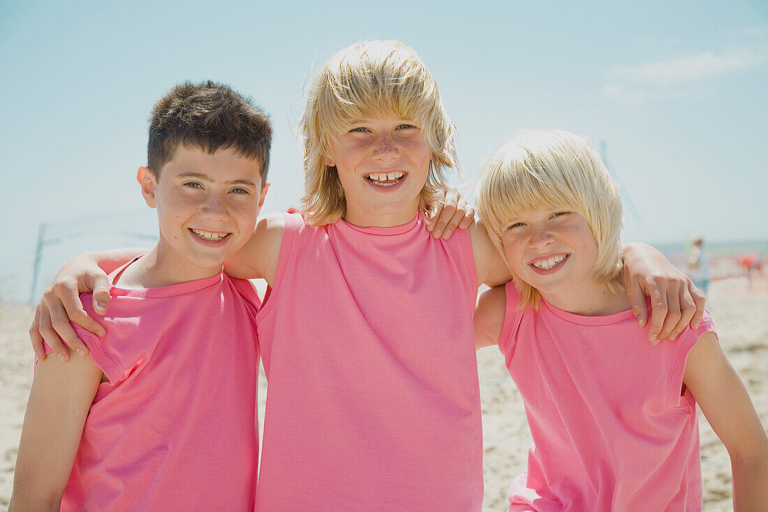 Three smiling boys on a beach wearing identical pink t-shirts\n