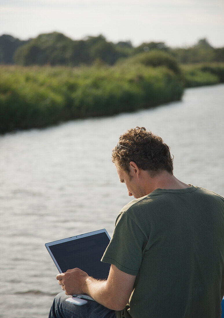 Rückenansicht eines Mannes, der an einem Fluss sitzt und einen Laptop benutzt