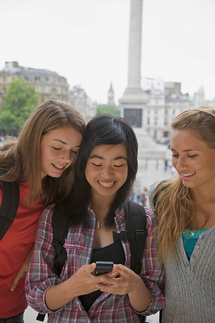 Drei lächelnde Mädchen im Teenageralter benutzen ein Handy am Londoner Trafalgar Square