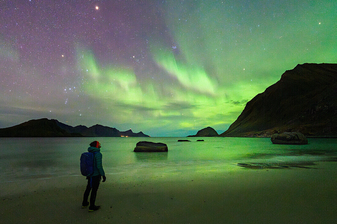 Mann mit Rucksack bewundert die hellen grünen Lichter der Aurora Borealis (Nordlichter) vom Haukland Strand, Lofoten Inseln, Nordland, Norwegen, Skandinavien, Europa