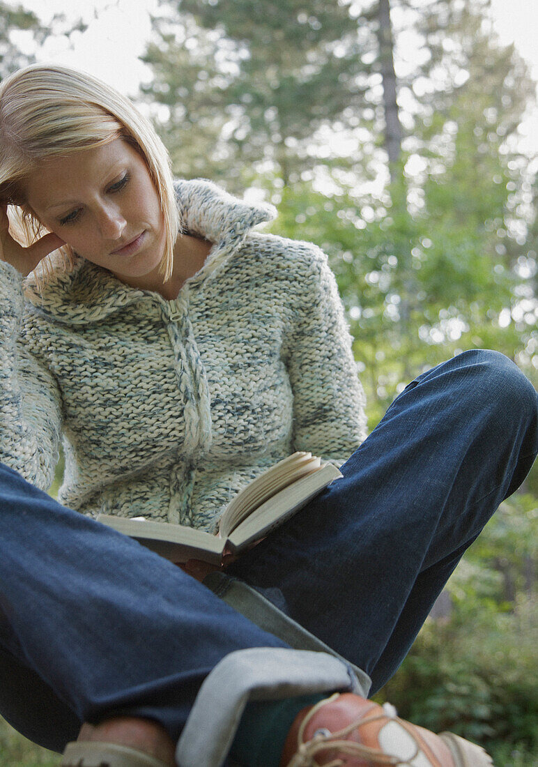 Junge Frau sitzt im Schneidersitz im Wald und liest ein Buch