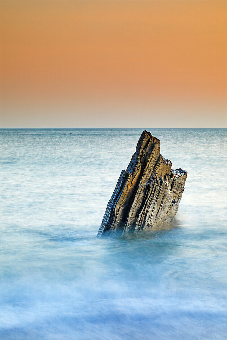A sunset view of a jagged tooth-like rock, surrounded by surging sea at Ayrmer Cove, a remote cove near Kingsbridge, south coast of Devon, England, United Kingdom, Europe\n
