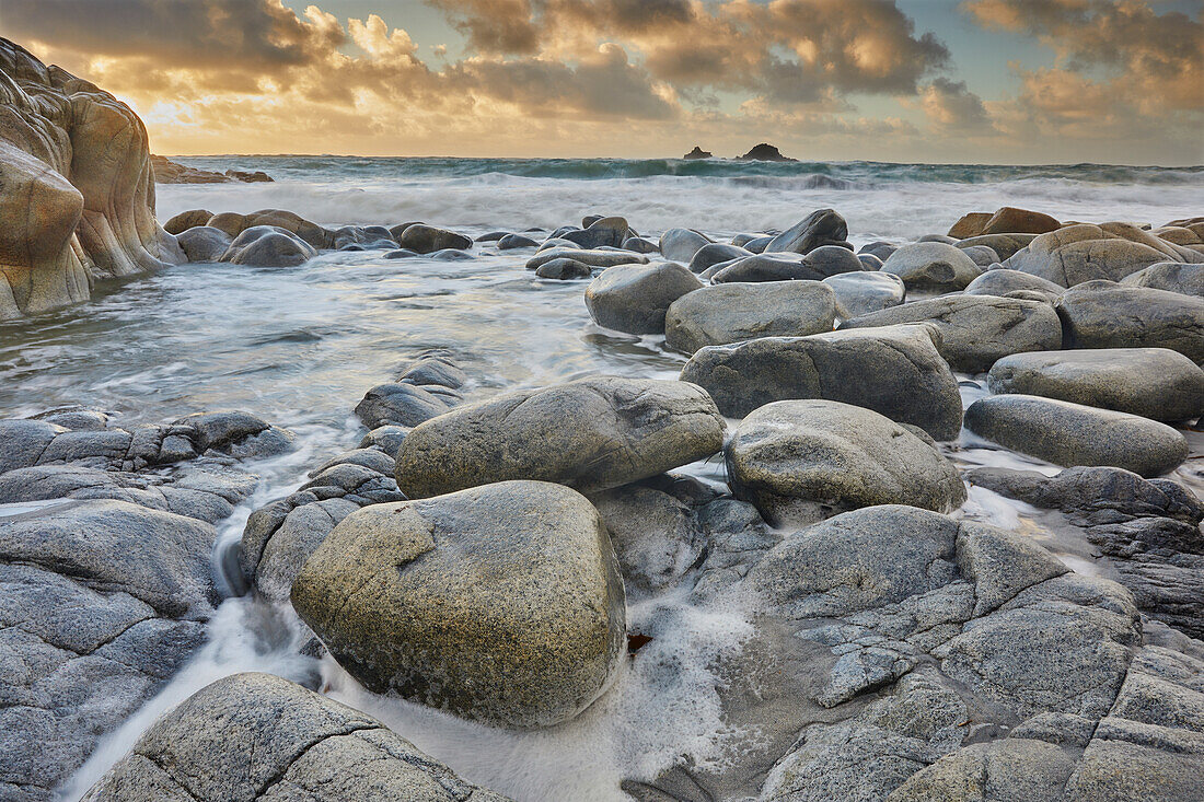 Atlantic rollers surge around shoreline granite boulders on a rising tide at sunset, Porth Nanven, a remote cove near St. Just, in the far west of Cornwall, England, United Kingdom, Europe\n