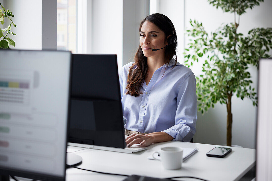 Lächelnde mittelgroße Geschäftsfrau mit Headset im Büro vor dem Computerbildschirm