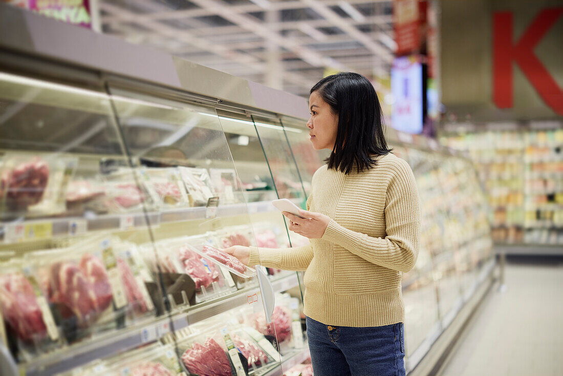 Frau kauft im Supermarkt ein und benutzt ihr Handy, um Preise zu vergleichen oder die Einkaufsliste zu prüfen