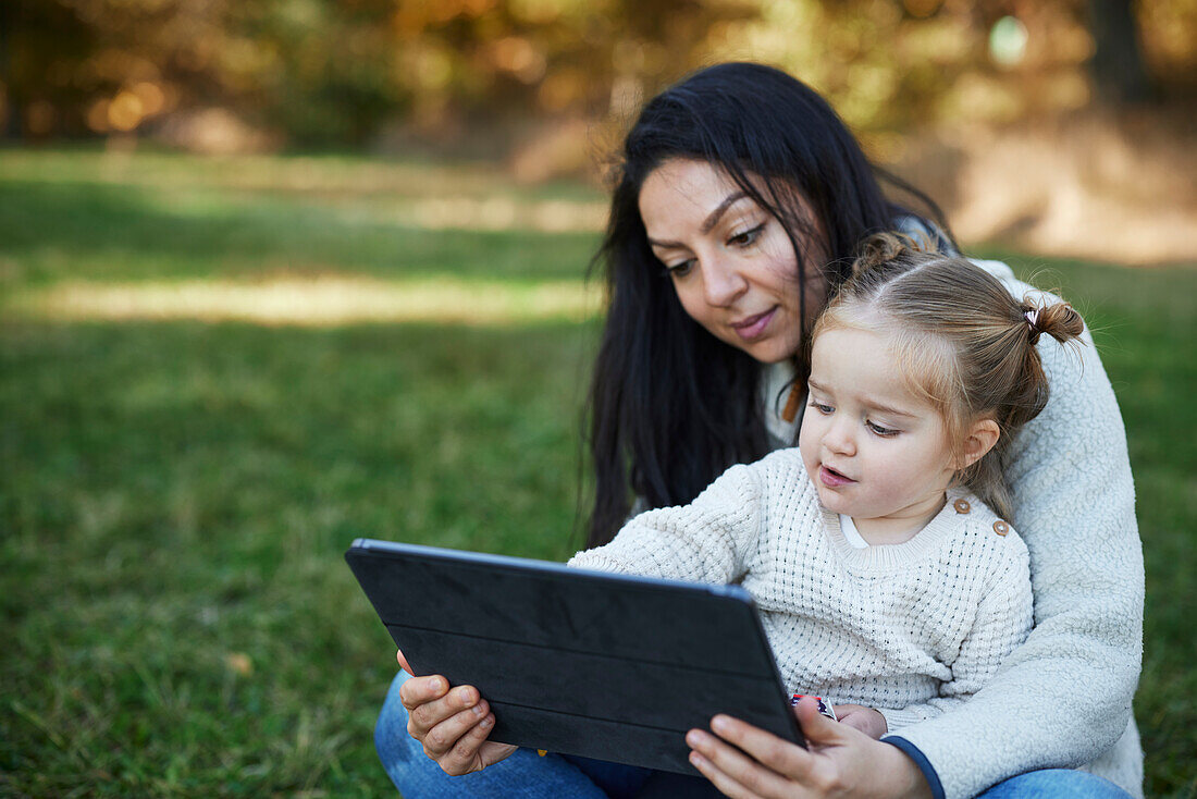 Mother and daughter sitting in autumn park and using digital tablet\n